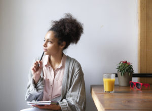 Image shows pensive-looking young biracial woman sitting at a table, which holds a full glass of orange juice and a pair of red-framed sunglasses, while she holds a pen to her face with one hand and a notebook with the other.