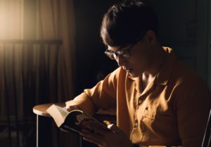 Image shows young Asian man sitting by a table and reading a book. A curtained window with some light shining through, and on his face, can be seen in the background.